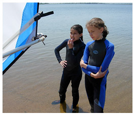 Julia and Anna wearing wet suits beside windsurf board.